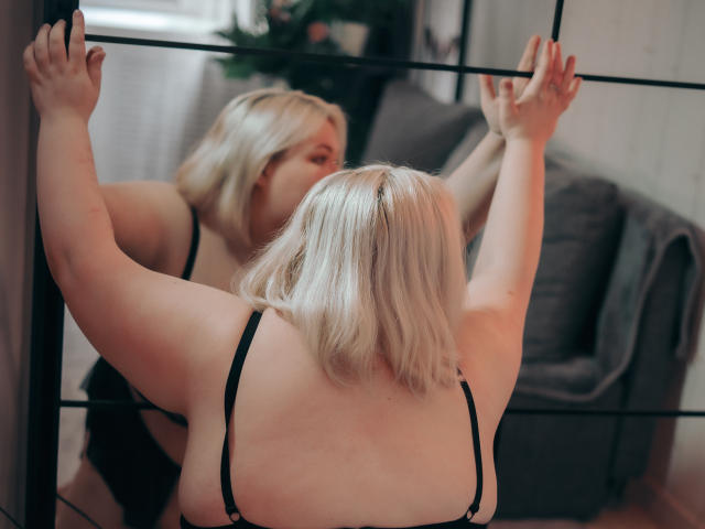 EllaBigBabe met kort blond haar, armen omhoog tegen een spiegel, draagt een zwarte beha, waarbij haar rug en borsten worden weergegeven, in een zacht verlichte kamer. Haar reflectie toont een enigszins nieuwsgierige uitdrukking..