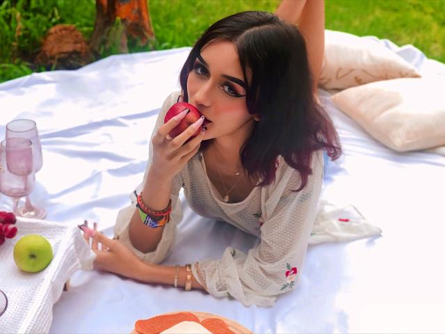 KhloeLopez, with purple-tipped black hair, bites a red apple, wearing a white dress, lying on a white blanket, surrounded by pink glasses, green apple, and orange slice.