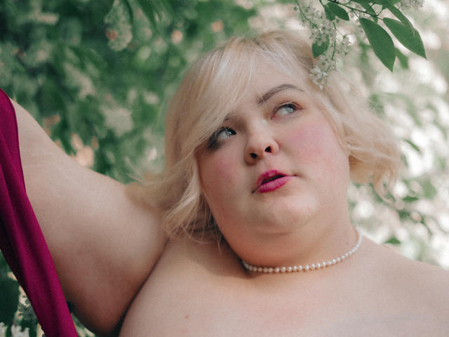 EllaBigBabe with blonde hair, pink lipstick, pearl necklace, and raised arm in a red garment, looking up with a slightly curious expression. Her face is chubby with pink cheeks.
