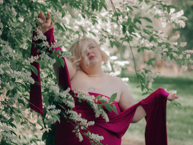 EllaBigBabe with blonde hair, pink dress, exposed boobs, arm raised, holding white flowers, green leaves, soft light, slightly blurry background, peaceful expression, outdoor setting.