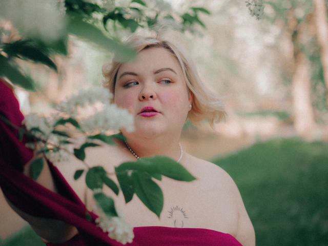 EllaBigBabe with blonde hair, pink lips, and a tattoo on her chest, wearing a red off-shoulder top, standing amidst green leaves and white flowers.