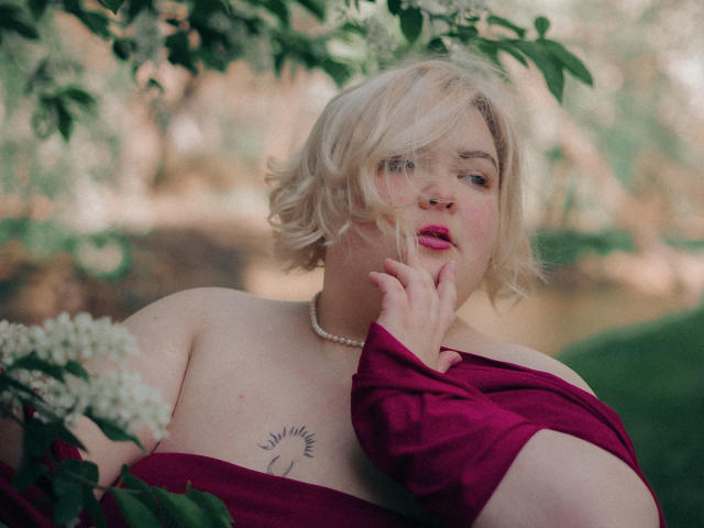 EllaBigBabe with short blonde hair, pink lips, and red dress, touching her chin, showing tattoo on chest, wearing pearl necklace, green leaves in background, soft sunlight.
