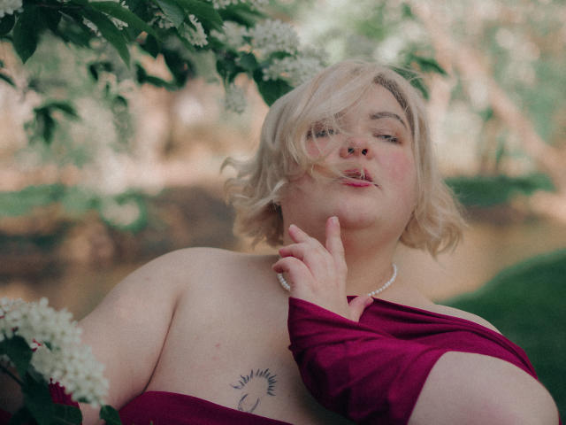 EllaBigBabe with blonde hair, pink lipstick, and a pearl necklace, wearing a red off-shoulder top, finger on chin, tattoo on right shoulder, surrounded by white flowers.