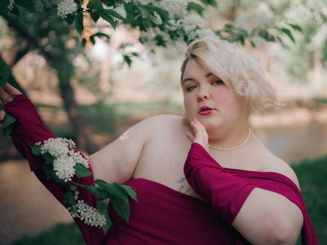 EllaBigBabe, chubby, with blonde bob, wearing off-shoulder magenta dress, pearl necklace, touching leaf, holding white blossoms, pink lipstick, looking at camera, soft light.