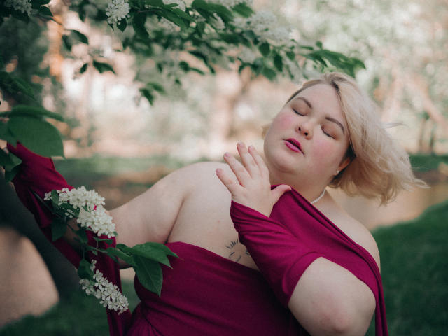 EllaBigBabe with her eyes closed, holding a magenta dress against her bare tits, surrounded by white flowers and green leaves, looking relaxed and sexy.