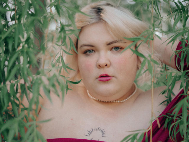 EllaBigBabe with blonde hair, blue eyes, pink lipstick, pearl necklace, red dress, and sun tattoo on chest, surrounded by green bamboo leaves. Her expression is serious and focused.