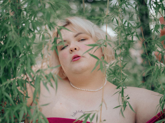 EllaBigBabe with blonde hair, pink lipstick, pearl necklace, and red dress, surrounded by green bamboo leaves, looking up with a slightly bored expression. Boobs partially visible.