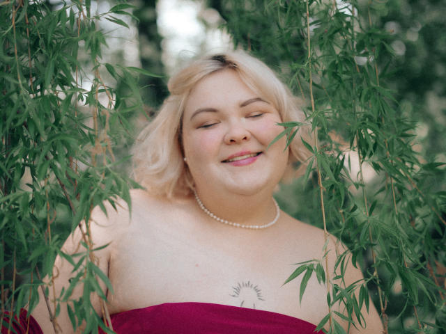 EllaBigBabe, blonde with a pearl necklace, smiling with eyes closed, wearing a red strapless dress, exposing sun tattoo on her chest, surrounded by green bamboo leaves.