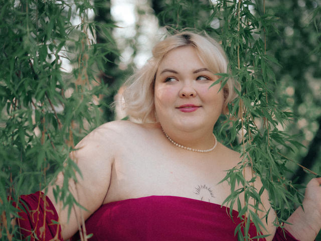 EllaBigBabe with blonde hair, red strapless dress, pearl necklace, smiling, green bamboo leaves in front, small sun tattoo on right chest, light skin, pink lips, medium build.
