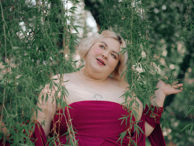 EllaBigBabe, chubby with pale skin, blonde hair, red dress, pearl necklace, hands in green bamboo leaves, slight smile, subtle makeup, small tattoo on chest, relaxed vibe.
