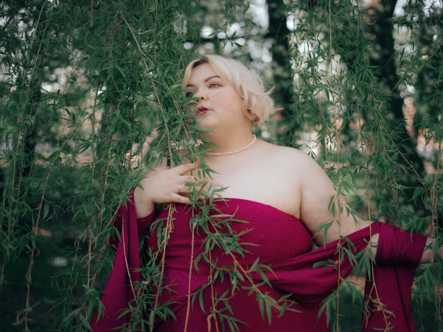 EllaBigBabe with pale skin, blonde hair, and a chubby build, wearing a red strapless dress, pearl necklace, and holding green leaves, standing in a forested area.