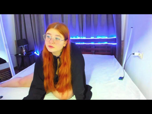 Emma: Uma mulher de cabelos ruivos, óculos e blusa preta, sentada em uma cama, mostrando sua bunda nua, olhando para a câmera, com uma fita de LED azul ao fundo..