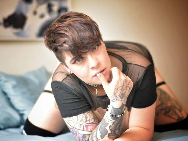 LenaaBlack with short brown hair, black sheer top, tattoos on arms and hands, black thigh-highs, finger near mouth, looking at camera, on blue bed.