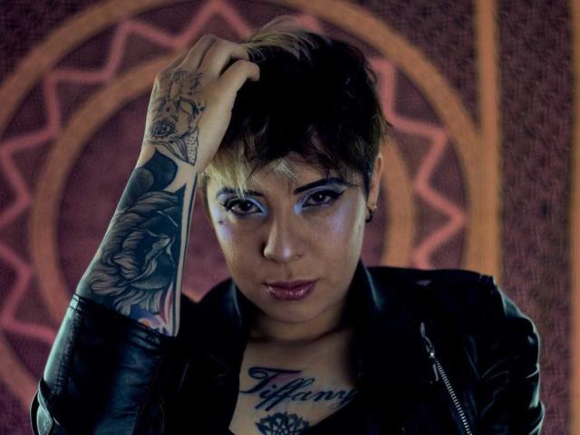 LenaaBlack with short brown hair, dark eye makeup, tattoos on left arm and chest, wearing black leather jacket, hand in hair, intense gaze, slightly parted lips.