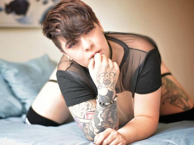 LenaaBlack, with short brown hair, black sheer top, and tattoos, kneels on a bed, sucking her finger, looking at the camera, showing off her black thigh-high socks.