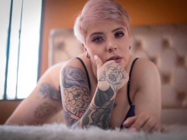 LenaaBlack with pink hair, tattoos on arms and hands, black bra, lying on bed, finger in mouth, looking at camera, soft light, orange wall, fluffy white blanket.
