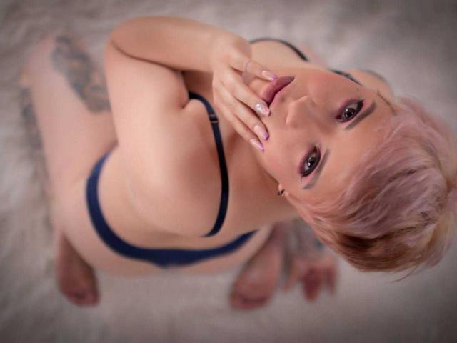 LenaaBlack with pink hair, black bra, and navy panties, looking up with hand near mouth, tattoos on thigh, sitting on fluffy white surface, showing her tits and ass.