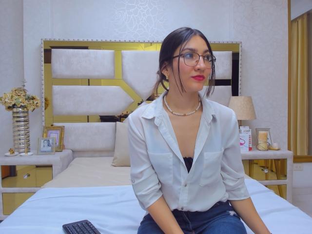 Selene Roussel, avec des lunettes, du rouge à lèvres rouge, une chemise blanche, un soutien-gorge noir, un collier de perles, assise sur un lit, en jean bleu, avec un clavier noir devant elle, dans une chambre avec une tête de lit jaune et des lampes..