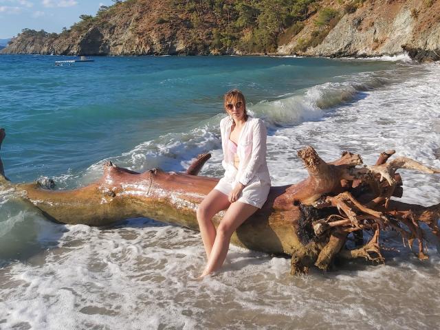 Sadie, con i capelli biondi e gli occhiali da sole, è seduta su un tronco bagnato e sbiancato dal sole, nell'oceano, indossando una camicia bianca a maniche lunghe con bottoni e un paio di pantaloncini da bagno rosa..