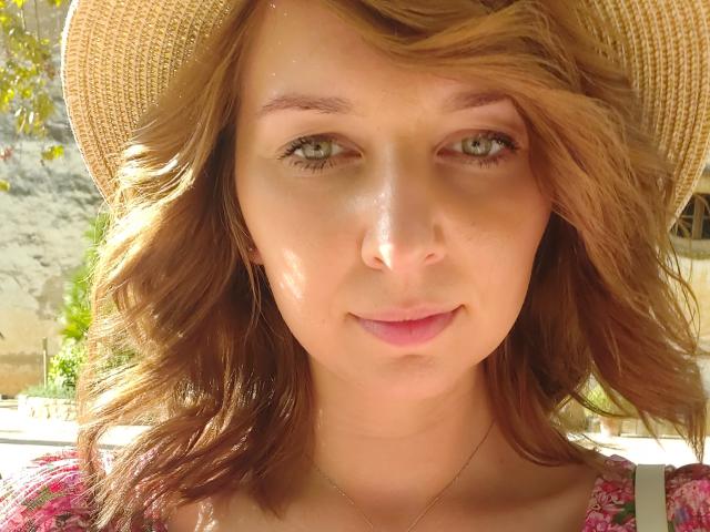Sadie Firt hat hellbraunes, welliges Haar, grüne Augen und ein leichtes Lächeln. Sie trägt einen Strohhut und ein pinkes Oberteil mit Blumenmuster, hat sonnengebräunte Haut und dezentes Make-up..