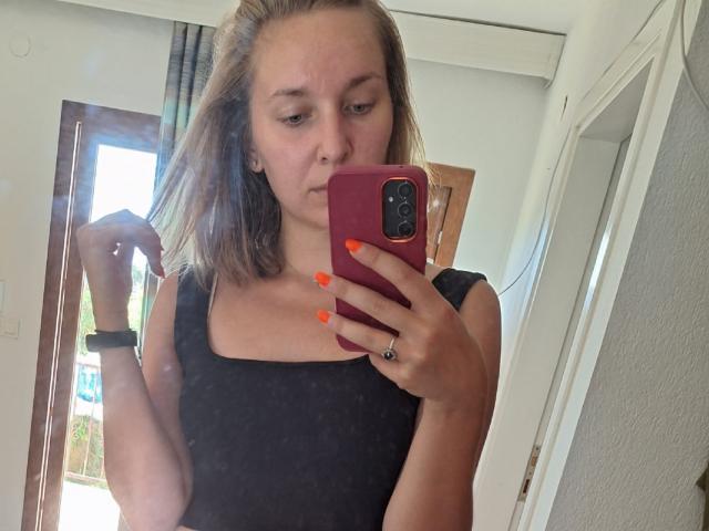 SadieFirt, met bruin haar dat tot op haar schouders reikt, draagt een zwarte top, houdt een roze telefoon vast met oranje nagels, trekt een frons en draagt een zwarte polsband, op een spiegel-selfie..
