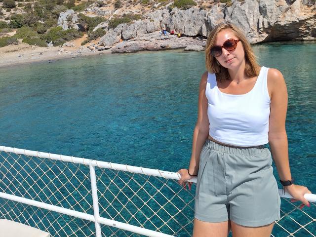 Sadie Firt mit blonden Haaren, einem weißen Tanktop und grauen Shorts, lehnt an einem Zaun, trägt eine Sonnenbrille und steht neben klarem, blauem Wasser, im Hintergrund ist ein felsiger Strand zu sehen..