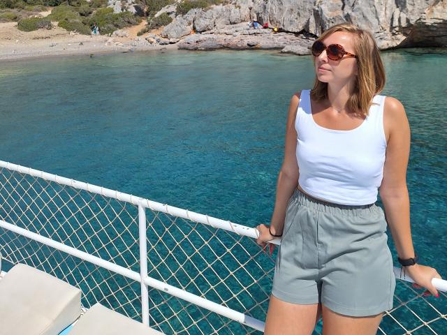 SadieFirt, avec une poitrine de taille moyenne, un débardeur blanc, un short gris, des cheveux bruns, des lunettes de soleil, debout près d'une rambarde en métal blanc, tenant un objet blanc, avec de l'eau bleu-vert en arrière-plan..