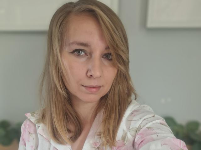 Sadie Firt ha i capelli biondi, gli occhi verdi e un nasino piccolo con un leggero sorriso. Indossa una camicia a fiori e guarda direttamente la fotocamera con un'espressione rilassata..