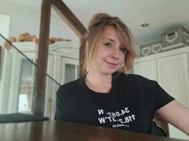 SadieFirt, loira, fofa, sorrindo, camiseta preta, cabelo curto, seios pequenos, pele clara, em pé, mesa de madeira, cerca de metal ao fundo, casual, interior, luz natural, relaxada..