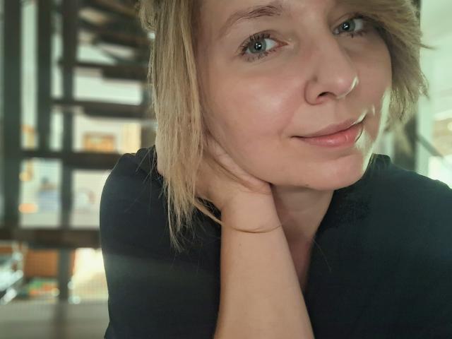 Il viso di SadieFirt, capelli biondi, occhi azzurri, leggero sorriso, maglietta nera, mento appoggiato sulla mano, luce soffusa, casual, carina, trucco naturale, bellezza delicata, rilassata, capelli leggermente spettinati..
