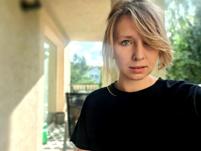 SadieFirt, met blond, golvend haar dat één oog bedekt, draagt een zwarte trui, heeft lichtgroene ogen en een neutrale uitdrukking, en staat in fel zonlicht..