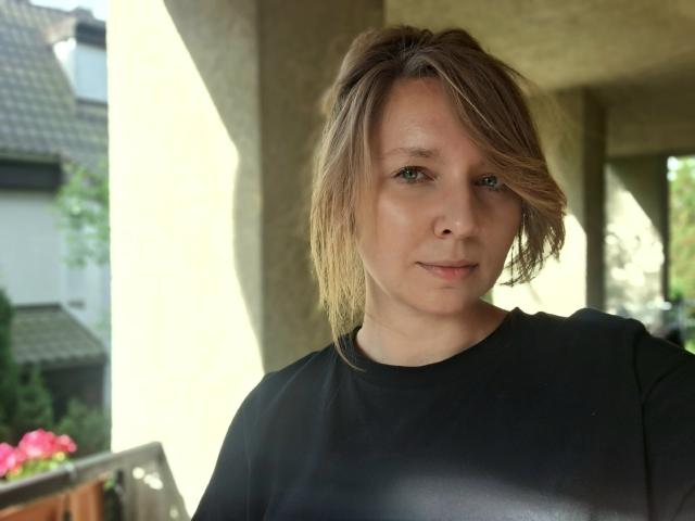 SadieFirt ha un leggero sorriso, indossa una maglietta nera, con i capelli biondi raccolti in modo disordinato, occhi verdi e pelle chiara, ed è in piedi in un'area illuminata dal sole, ma in ombra..