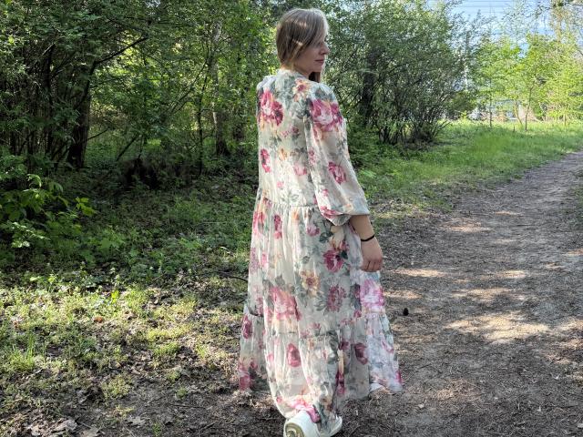 SadieFirt met blond haar, een bloemenjurk, met de rug naar de camera, staand op een zandpad, kijkend over haar schouder, met groene begroeiing eromheen, witte sneakers, een lichte glimlach, een zomerdag, de losse jurk wappert in de wind..