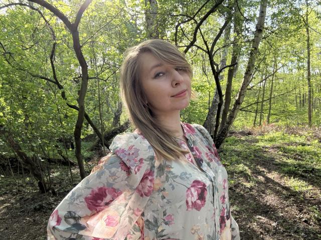 Sadie Firt, met blond haar, een bloemenjurk en roze rozen, kijkt op met een lichte glimlach in een zonnig bos. Ze heeft een schattig gezicht en kleine borsten..