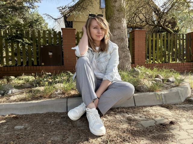SadieFirt zit op een betonnen rand, gekleed in een denimjas, grijze broek, witte sneakers en met een zonnebril op haar hoofd. Ze heeft blond haar en een lichte glimlach..