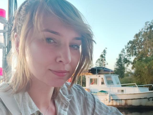 SadieFirt, con i capelli biondi, la pelle chiara e le labbra rosa, indossa una camicia a righe, sorride leggermente, con una barca e degli alberi sullo sfondo, in una giornata di sole..