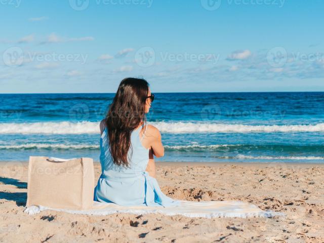 DaliaForFun, con el pelo largo y castaño, vestida con un vestido azul, está sentada en una playa arenosa, mirando hacia el océano, con una bolsa de arena a su izquierda y gafas de sol..