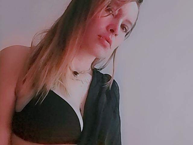 LillyThompson with pinkish-red face, pouty lips, and half-shaded eyes, wearing a black bra and sheer black top, showing her small tits and blonde hair.