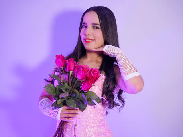 AlanaReyes with long black hair, pink lace corset, white fishnet gloves, holding pink roses, smiling with red lipstick, soft purple lighting, holding bouquet against white wall.