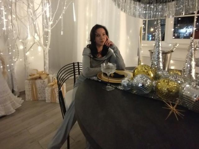 LexyTease with tits down, black hair, grey sweater, sitting at black table, Christmas ornaments, gift boxes, fairy lights, hand on chin, wooden floor, night, window, silver curtain.