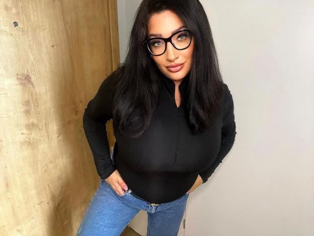 CindyQueen-hot with huge tits, black long-sleeve shirt, blue jeans, hands on hips, black glasses, long black hair, light skin, standing, confident, slightly leaning forward.