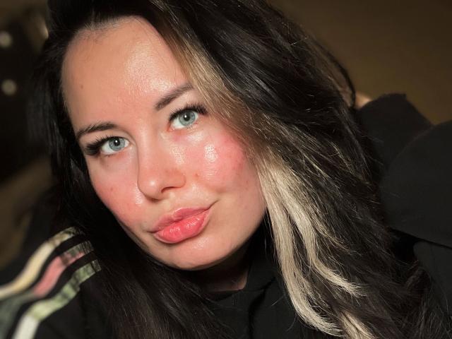Shyla, mit vollen, rosafarbenen Lippen, blauen Augen und schwarz-blondem Haar, blickt leicht schmollend in die Kamera und trägt ein schwarzes Hemd..
