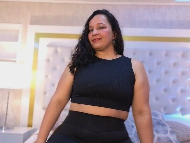 AmandaXBrown with black wavy hair, wearing a black ribbed sports bra and black high-waisted pants, smiling in a bedroom with a tufted headboard.