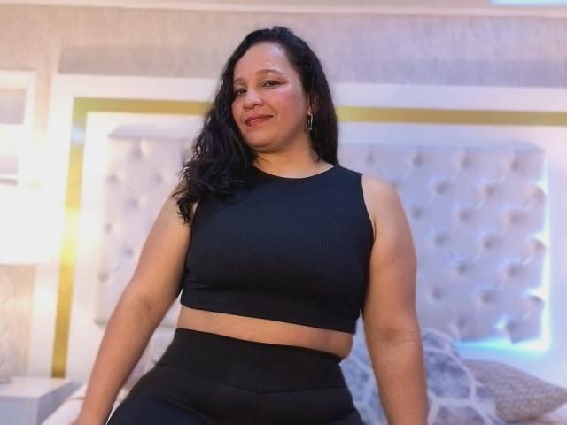 AmandaXBrown, eine kurvige Frau mit mittelbrauner Haut und schwarzen, welligen Haaren, trägt einen schwarzen Sport-BH und schwarze, hochgeschnittene Hosen und lächelt in einem sanft beleuchteten Schlafzimmer..