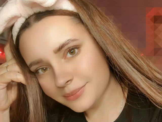 TiffanyKeli met lichtbruin haar, groene ogen en een lichte glimlach, draagt een roze hoofdband en raakt haar hoofd aan met een ring om haar vinger, in zacht licht..