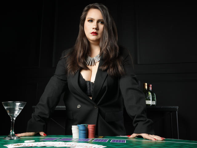 AvienLume with big tits, wearing black blazer and lace bra, leaning on green casino table, stack of chips, martini glass, and bottles in background, looking serious.