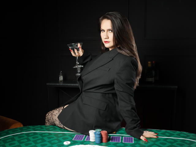 AvienLume with dark hair, wearing a black suit, holding a martini glass, sitting on a green poker table with chips and cards, showing her lace-patterned thigh.