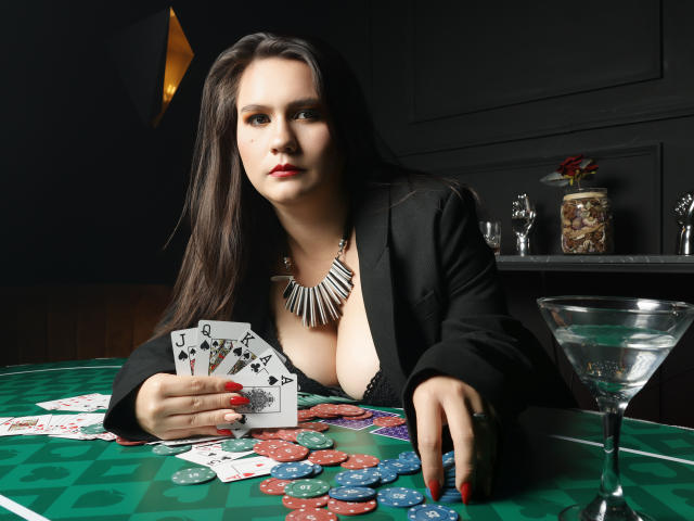 AvienLume with large boobs, in a black jacket, holding cards, and staring at a poker table with chips and a martini glass, has long dark hair and red nails.