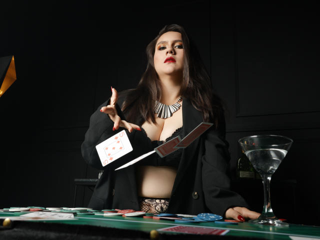 AvienLume with big tits in black lace, holding a card, sword in hand, over poker table with chips and martini, wearing black jacket and necklace, dark background.