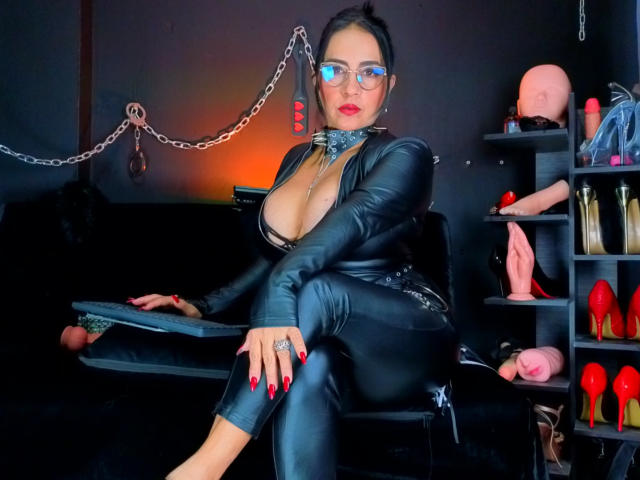 ScarletteStones in a black leather outfit, glasses, and red nails, sitting with a chain in the background, exposing her large tits and wearing a collar, holding a paddle.